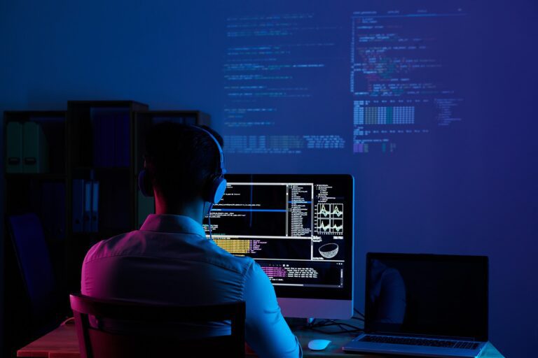 Osoba se sluchátky na uších sedí u stolu s počítačovým monitorem, na kterém se zobrazují kódy a data, a vedle ní je notebook. Počítačový kód je promítán na stěnu v tlumeně osvětlené místnosti.